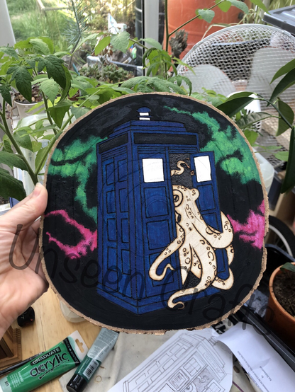 Oval wood slice with burned design showing an octopus coming out of a blue painted police call box with black background with vibrant green and pink streaks.