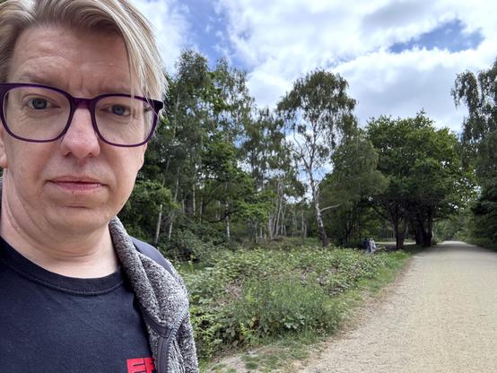 The author, a white middle-aged man with light blonde hair and  glasses with purple frames, looks into the camera with a pathway leading into the distance towards some trees. The sky has fluffy white clouds.