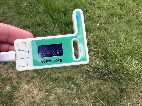 A small rectangular device in a white plastic case with a green label and an OLED display that cannot be read properly (it is corrupted due to the camera shutter speed). The device has a protrusion to the right side like an aerial. Text below the screen shows the URL heltec.org