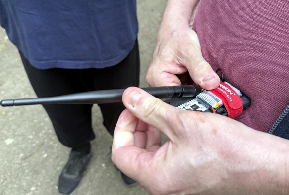 A pair of white-skinned hands holding a black aerial and a white circuit board with a red velcro strap.