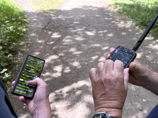 Two men out of shot hold wireless devices, on the left, an Android phone with colourful display that appears to show a list of items but is blurred, on the right, a walkie-talkie like device with a black aerial. Hands are operating the devices. A pathway leads into the background of the image.
