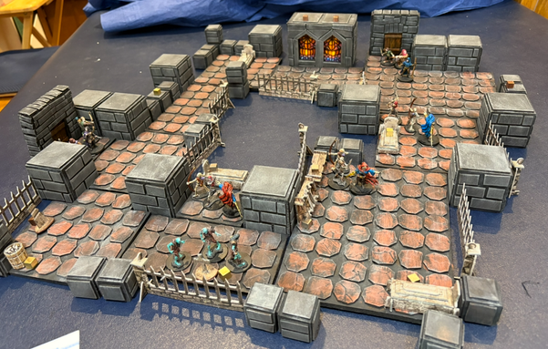 Photo of a tabletop game setup showing a dungeon map with a roughly circular shape. Various undead enemies are sprinkled around the map and two groups of adventuring heroes are entering the map at the far end and on the left.