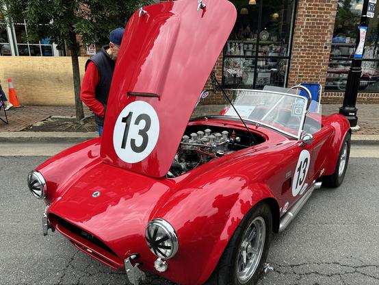 The image features a classic red sports car with its hood open, revealing the engine. The car is adorned with the number "13" on both the hood and the side, indicating it may be a race car. The vehicle has a sleek, aerodynamic design with a convertible top, showcasing its vintage appeal. The engine compartment is visible, with multiple chrome-plated components and a polished appearance. The car is parked on a street, with a brick building and a window display in the background. A person wearing a red shirt and a blue cap is standing next to the car, seemingly inspecting it. The scene is set in a public area, possibly during a car show or exhibition.

Provided by @altbot@fuzzies.wtf, generated privately and locally using Ovis2-8B

🌱 Energy used: 0.171 Wh