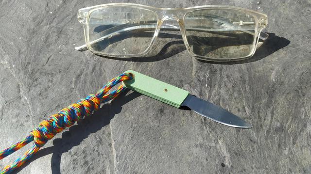 A pair of glasses (for scale)  and a tiny knife with 3D printed scales and a paracord lanyard sat on a slate slab.