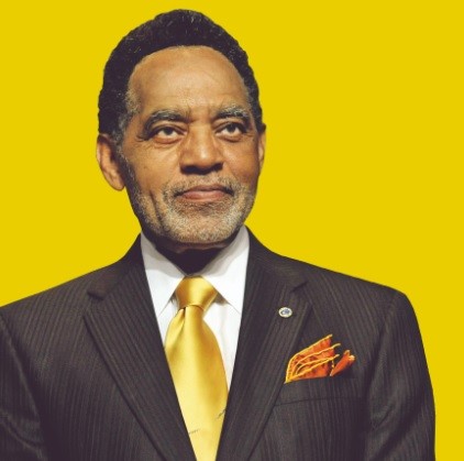 Burrus in a suit, with yellow tie, against a yellow background, with graying, very trim beard, looking straight ahead (eye contact).
