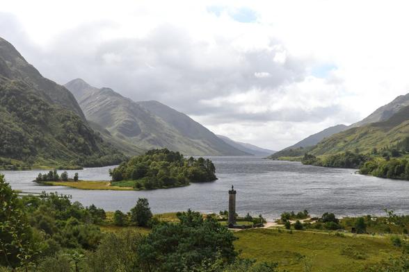 Malerisch zwischen Berghängen in einem engen Tal liegt ein See, darin nahe dem Ufer eine bewaldetes Inselchen (Eilean Ghleann Fhionainn). Am Ufer steht eine gemauerte Säule, auf deren Kopf eine Figur steht – das Glenfinnan-Denkmal, das den Soldaten aus dem schottischen Hochland im Jakobiteraufstand 1745 gewidmet ist. Menschen laufen am Ufer herum. Es ist sonnig aber bewölkt.