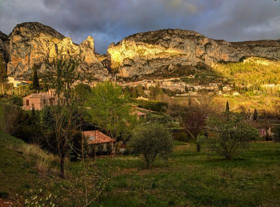 A wide shot captures the picturesque village of Moustières-Sainte-Marie nestled at the base of large, rocky cliffs under a dramatic sky. The cliffs, illuminated by a warm, golden light, dominate the upper portion of the frame, revealing their textured, layered surfaces. Below, the village consists of closely packed buildings with terracotta roofs, partially obscured by lush green trees and vegetation. The foreground features a grassy area dotted with various trees and shrubs, some showing fresh spring foliage. The sky above is a mix of dark, brooding clouds and patches of lighter sky, suggesting either an approaching or receding storm, with the sunlight breaking through to highlight parts of the landscape. The overall mood is one of serene beauty, contrasting the ruggedness of the cliffs with the quaintness of the village.