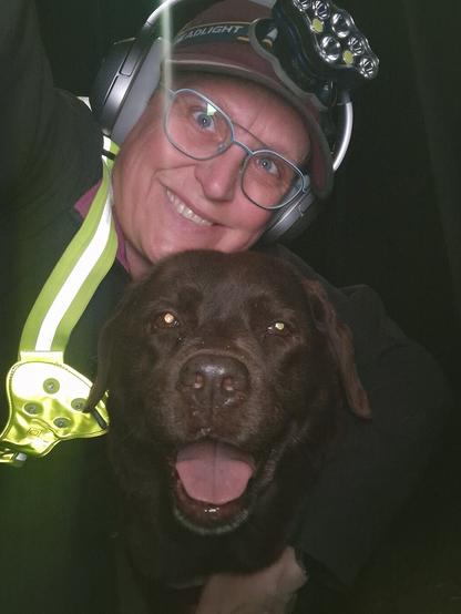 Night time selfie of a person and a chocolate brown Labrador with greying snout. The person is wearing a black jacket, yellow safety harness, red cap, silver headphones and blue-rimmed glasses. On their cap rests a headstrap torch in off position. Their free hand is around the dog's shoulders. Both are facing the camera. The dog's mouth is slightly open, showing off some mlem. The person's head is resting on the dog's head. They're smiling, grateful for their time spent together.