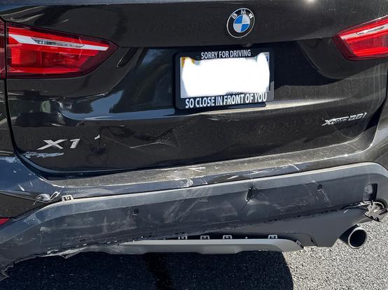 The image shows the rear of a black BMW X1 with visible damage to the rear bumper. The car's license plate is obscured, but a sticker on it reads "SORRY FOR DRIVING SO CLOSE IN FRONT OF YOU." The BMW logo is prominently displayed at the top center of the trunk. The rear bumper shows significant damage, with dents and scratches, particularly on the right side. The car's model designation "X1" is visible on the left side of the trunk, and "XDrive 25i" is on the right. The taillights are intact, and the car appears to be parked on a paved surface.

Provided by @altbot@fuzzies.wtf, generated privately and locally using Ovis2-8B

🌱 Energy used: 0.156 Wh