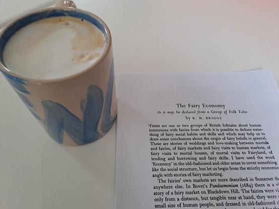 Photo of a coffee mug with coffee and milk foam in it, and the printed pages of an article titled "The Fairy Economy, as it may be deduced from some folk tales" by K. M. Briggs