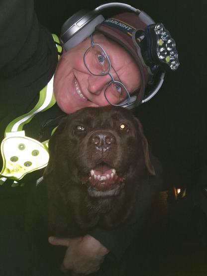Night time selfie of a person and a chocolate brown Labrador with greying snout. The person is wearing a black jacket, yellow safety harness, red cap, silver headphones and blue-rimmed glasses. On their cap rests a headstrap torch in off position. Their free hand is around the dog's shoulders. Both are facing the camera. The dog's mouth is slightly open, showing off some mlem. The person's head is resting on the dog's head. They're smiling, grateful for their time spent together.