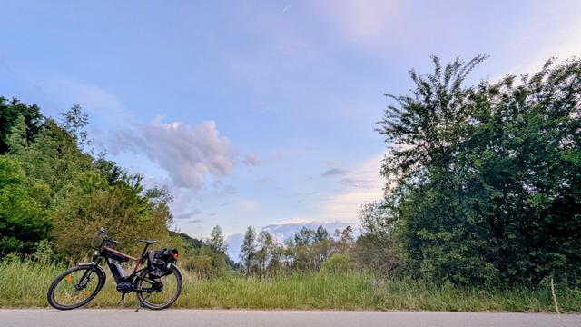 Das Bild zeigt ein idyllisches Naturpanorama am frühen Morgen. Im Vordergrund steht ein modernes E-Bike am Straßenrand, leicht schräg auf einem ruhigen Asphaltweg geparkt.

Im Hintergrund erstreckt sich eine dichte, grüne Vegetation mit verschiedenen Bäumen und Sträuchern. Der Himmel ist größtenteils klar, mit wenigen, leicht rosa gefärbten Wolken, die goldene Stunde. Die Farben des Himmels reichen von sanftem Blau bis zu pastellfarbenem Rosa und Gelb