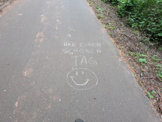 Ein asphaltierter Wanderweg auf dem mit Kreide der Spruch "Hab einen schönen Tag" ausgemalt ist. Unter dem Spruch ist noch ein lächelndes Smiley gemalt.