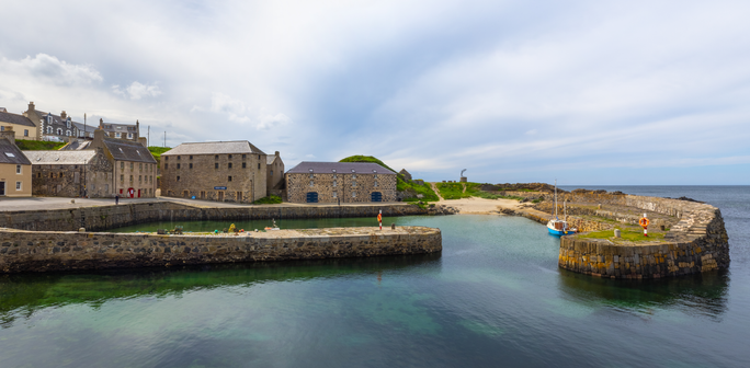 A wall art piece featuring a 17th century Scottish Harbour, the ancient buildings stand timelessly as the back drop to the ancient thick stone harbour walls a light cloud sky reflects in the still waters with gentle calm reflections.