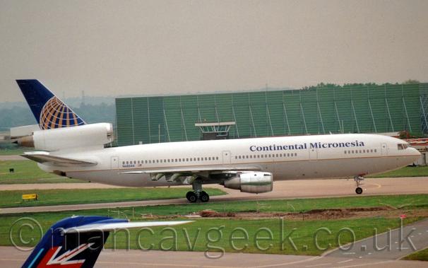 Side view of a white, 3 engined jet airliner taxiing from left to right.
The plane is mostly white, with a grey lower half, with gold piping separating the 2 sections, and dark blue "Continental Micronesia" titles on the upper forward fuselage.
Large patches of green grass in the foreground separate different taxiways.
A large, green, box-like metal structure stretches across the frame from the right, under grim grey skies.