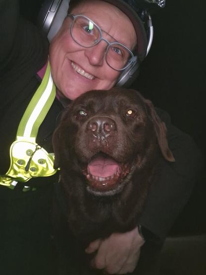 Night time selfie of a person and a chocolate brown Labrador with greying snout. The person is wearing a black jacket, yellow safety harness, red cap, silver headphones and blue-rimmed glasses. On their cap rests a headstrap torch in off position. Their free hand is around the dog's shoulders. Both are facing the camera. The dog's mouth is slightly open, showing off some mlem. The person's head is resting on the dog's head. They're smiling, grateful for their time spent together.