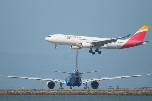 An Iberia A330 twin engine aircraft is landing over a runway with a blue ITA A330-9 plane positioned in the foreground, lined up and ready for takeoff. The background is a hazy sky. The foreground has calm San Francisco bay waters.