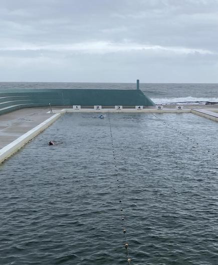 Looking over the lap pool and out to sea. A couple of swimmers are in lanes 1 and 2. Grey skies and ocean behind the curved turquoise concrete bleachers and wall.