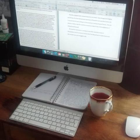 A screen showing two opened documents of text and its wireless keyboard and mouse on a wooden desk, between them a fibre-tipped pen upon a loose-leaf book open at two pages of handwritten notes, and beside the book a drink of raspberry tea in a white china cup decorated with a golden rim.