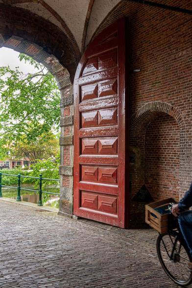 A gate with high doors that are open so that cyclists can pass through. From the left below the middle, the street runs diagonally. Then above that, about halfway to the left, the opening of the gate, behind that you see green leaves of trees through the gate and a small gate of a bridge. Next to it, the high, brown-red painted door with an arch at the top. Next to it, the old stone wall of the gate. On the right, you see the wheel of a cyclist who is coming and who has just come through the gate on the other side.