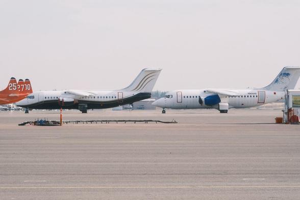 Two BAE-146 airplanes parked on an airport tarmac. The plane on the left is white with black and orange accents, while the plane on the right is white with partially scraped off blue accents. Three orange-red CalFire aircraft with numbers painted on their tails are partially visible.