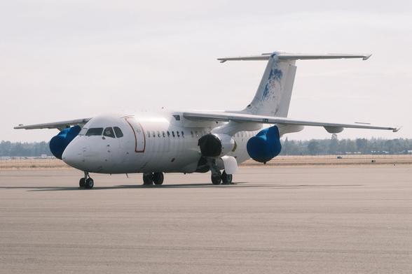 A parked four engine BAE-146 on an airport tarmac with engine covers on. The aircraft with poorly scraped off blue accents has a high-wing design and a distinctive T-tail. Its door is taped off with red tape. The background has a blurred hazy flat landscape with trees.