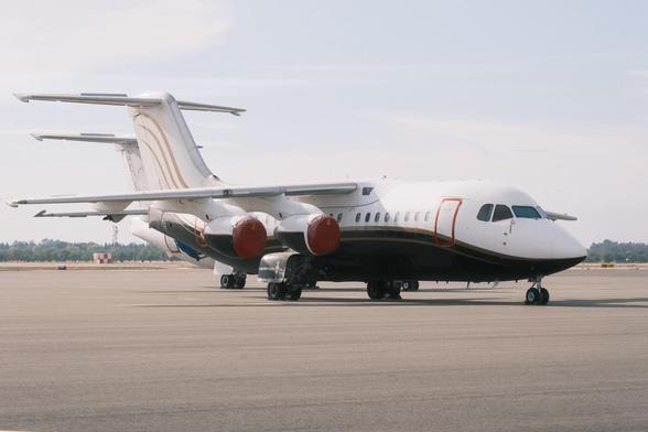 A profile view of a parked BAE-146 on an airport tarmac with four engines, distinctive red engine covers, and a T-tail design. The fuselage is white with black and gold accents. A second BAE-146 is behind the first one and is barely visible.