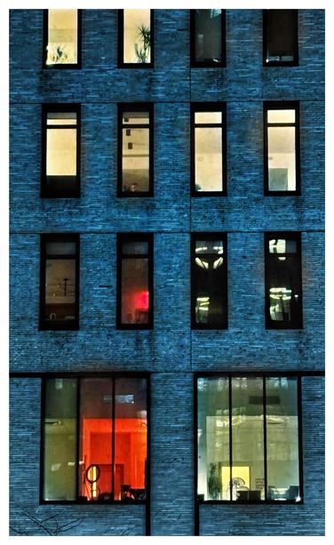 Photo of tall, narrow rectangular windows with lit interiors in a building wall. The facade is a dark shade of blue and is made of narrow horizontal bricks which give it a textured look. On the lowest level are two larger windows, each divided vertically into three panels. Above them are three more levels with four smaller windows each; the upper windows are cut off at the top of the image. The interiors seen through the windows are variously lit in tones of red and pale green, and show plants or doorways or bare walls or reflections of the city outside.