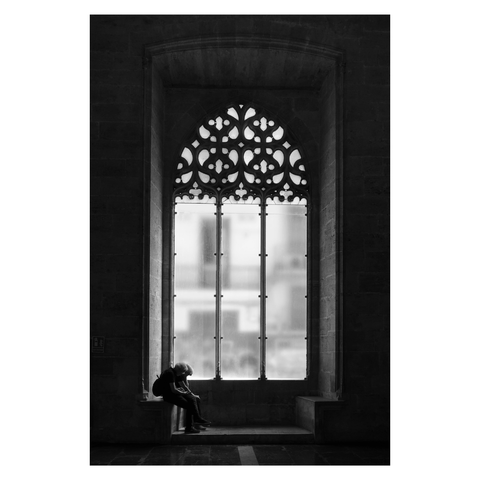 This is a portrait format black and white image of a large medieval window. Two people sit in a seat set below the window. Light falls through the window, picking out the details of the stonework. The interior is dark, and the two figures are almost in silhouette. Taken in the Silk Exchange, Valencia, Spain.