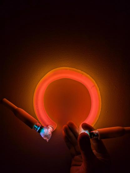 A pink and gold horseshoe of light held against a white wall so that the inside of the horseshoe is pink and the outside is gold with argon mercury blue, blackened electrodes and boots coming out the front ends of the glass tube