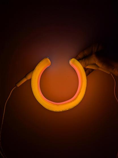 A pink and gold horseshoe of light held against a white wall so that the inside of the horseshoe is pink and the outside is gold. A hand is holding the tube from behind.