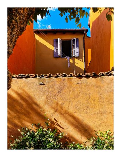 The photo is of a yellow stucco house with a window on the right which has open purple shutters. There is a washing line attached to it and hanging on it is a purplish garment is hanging to dry. The house is surrounded by a low wall, also made of yellow stucco, topped with terracotta tiles. A tree stands on the left side of the image, casting shadows on the wall and the house. In the foreground, green plants with small white flowers are visible at the bottom of the image. The background showcases a clear blue sky with a few white clouds.