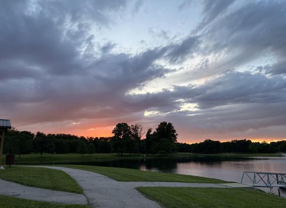 Scenic view of a park at sunset, featuring a tranquil lake, lush green grass, and a tree-lined horizon under colorful clouds. A curved pathway leads towards the water.
