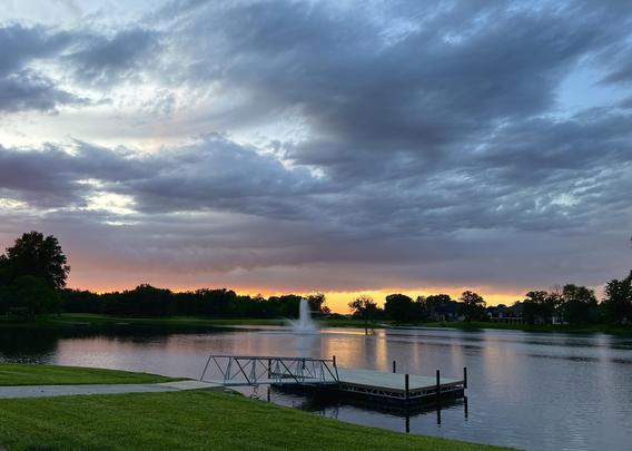 A tranquil lake scene at sunset, featuring a wooden dock in the foreground, a fountain in the water, and dark, cloud-filled skies above. The horizon glows with soft hues of orange and purple.