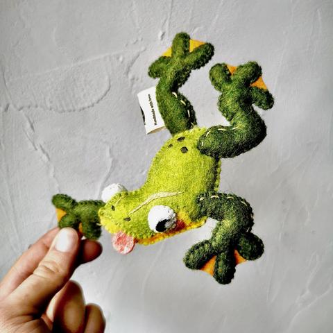 A green frog plushie held in front of a white wall. 
The frog is faceing downward, as if mid-jump. Its body is a bright green and it’s belly yellow, it’s pink tongue sticking out a bit. 
It’s four legs are a dark green with orange webbing between it’s round toes.
