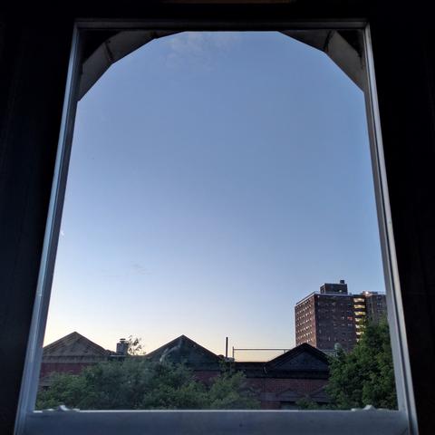Looking through an arched window twenty-one minutes before sunrise the sky fades from white at the horizon to pale blue above, with a small wisp of cloud high up in the sky. Pointed roofs of Harlem brownstones are silhouetted across the street, and a taller apartment building can be seen in the distance. The tops of two trees are in the shadows on the bottom and right.