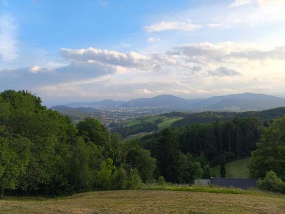 Im Vordergrund Wiesen und Wälder. Blick hinunter auf eine Großstadt. Dahinter einige Hügel. Blauer Himmel mit ein paar Wolken.