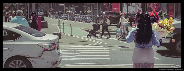 Two people stand across the street from one another, one raising a phone to snap a picture of a bouquet of flowers that the other is holding.  Other pedestrians and cars are visible throughout.