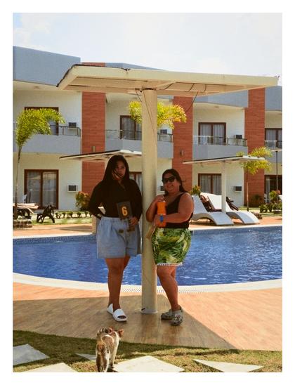 My godmother (showing our table number) and mom (holding a Royal bottle) between the pool umbrella, with a cat at the bottom of the picture, walking towards them as they take a picture from my phone. There is a swimming pool and an accommodation building behind them.
