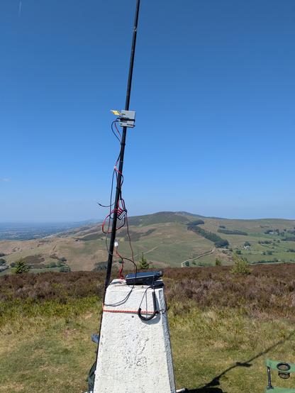 A trig point (painted white) has a fibreglass mast tied to it, with a radio sat on the top of the trig point. A messy run of cables runs up the mast to a metal box, with a yellow antenna attached to it. This is the SG-lab transverter with 2 element PCB antenna. The transverter is held to the mast using a velcro strap. This is as dodgy as it sounds.