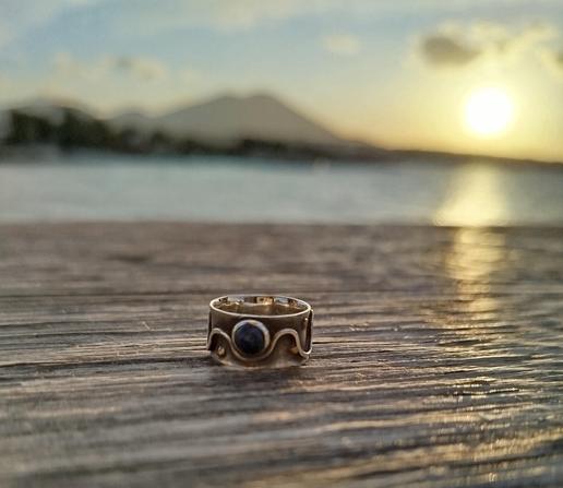 a wide silver ring with a small blue stone, the band has abstract wavy motifs. it's placed on a piece of wood. a sunset on background