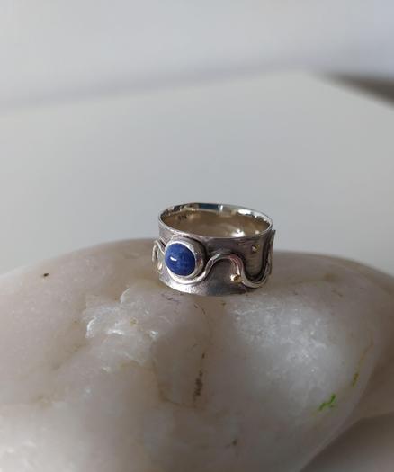 a wide silver ring with a small blue stone, the band has abstract wavy motifs. it's placed on a white pebble
