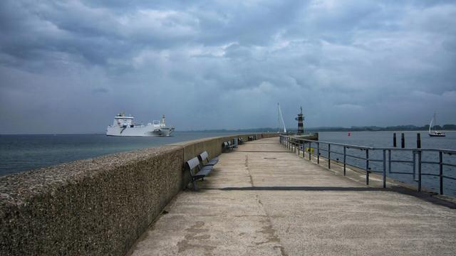 A long, grey concrete pier extends into a grey sea under a cloudy sky. Benches line the left side of the pier. A large white ship is visible in the distance on the left, and a black and white lighthouse stands at the end of the pier. A metal railing runs along the right side of the pier. The water is calm with a few buoys and a sailboat in the distance on the right.