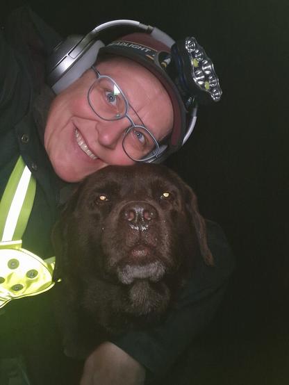 Night time selfie of a person and a chocolate brown Labrador with greying snout. The person is wearing a black jacket, yellow safety harness, red cap, silver headphones and blue-rimmed glasses. On their cap rests a headstrap torch in off position. Their free hand is around the dog's shoulders. Both are facing the camera. The person's head is resting on the dog's head. They're smiling, grateful for their time spent together.