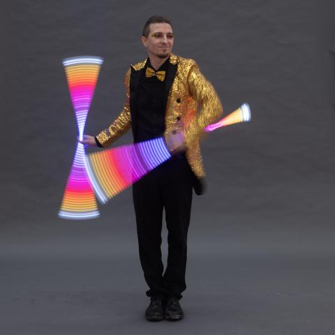 A performer in a glittery gold jacket and bow tie is skillfully juggling neon-colored light props. The background is a simple gray, emphasizing the vibrant colors of the juggling items.