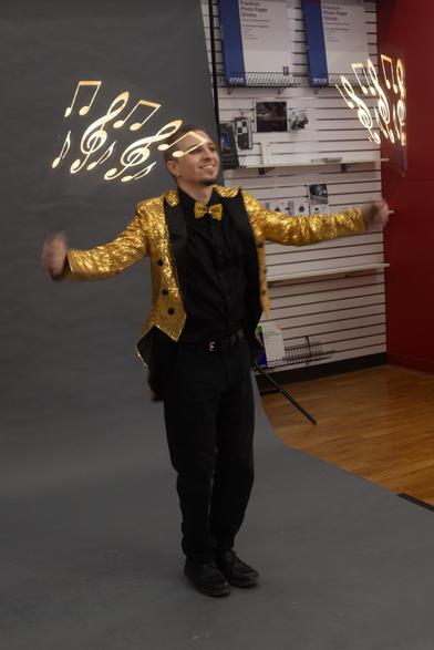 A person wearing a glittery gold jacket and bowtie performs energetically against a gray backdrop, with musical notes appearing above their head, suggesting a lively atmosphere. The setting includes shelves displaying various items in the background.