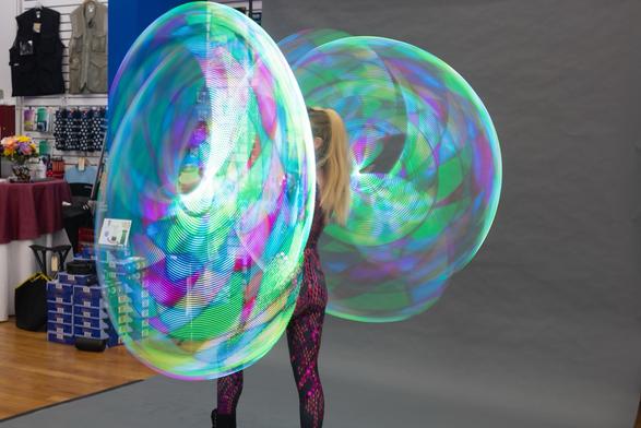 A person performs with four brightly lit, spinning hula hoops, creating colorful light patterns against a neutral background. Various objects and photography supplies are visible in the background.