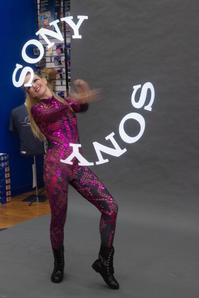 A performer in a shiny, pink sequined outfit poses dynamically against a gray backdrop. The word "SONY" is displayed in a circular motion around them, LED sticks being spun by the performer. The background includes various items related to photography.