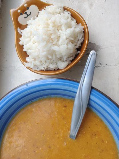 Blue bowl filled with orange dal, beside smaller brown bowl (with cat/bear face on the rim) with cooked white rice.