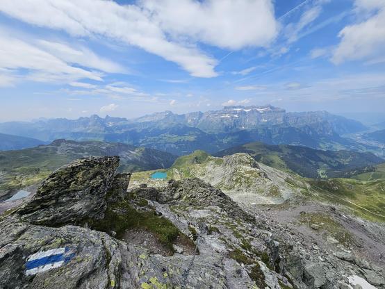 Foto einer Berglandschaft. Im Vordergrund Felsen und eine weiß-blau-weiße Markierung eines Alpinwanderwegs, in der Bildmitte ein türkisblauer Bergsee weiter unten und im Hintergrund ein Bergmassiv.